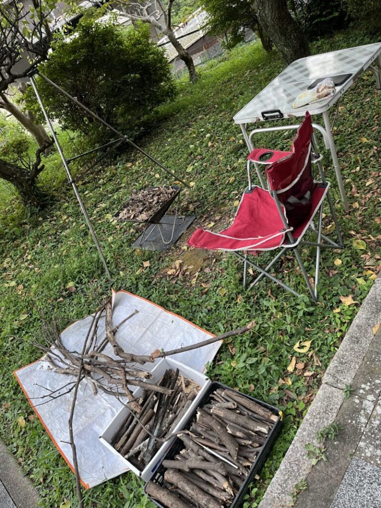 週末の焚き火の様子。焚き火台にトライポッドをセット。裏山から入手した枯れ枝と以前用意した薪が燃料。椅子と机を用意し、いつでも開始可能。