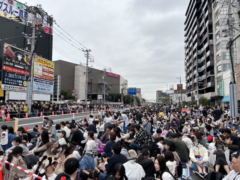 黒崎ディズニーパレードを待つ大勢の人々が沿道に並び、カメラを構えたり手を振ったりしながら、ミッキーとミニーの登場を心待ちにしている様子。色とりどりの服を着た観客が笑顔で集まり、街全体が祝祭ムードに包まれている。