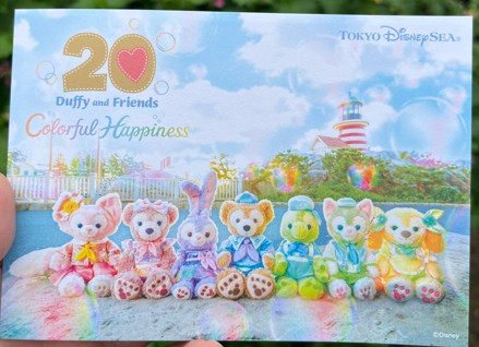 長浦公園で配布されたダッフィー20周年記念パンフレットの写真。表紙にはダッフィーと仲間たちが虹とシャボン玉に囲まれて微笑んでいるイラストが描かれ、優しい色合いとふんわりしたデザインが来場者の注目を集めている。パンフレットを手にした人々が周囲に集まり、記念に持ち帰ろうとする様子も見られる。