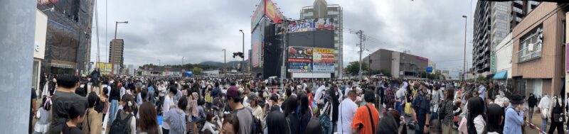 ディズニーパレード終了直後の黒崎の道路に、余韻を楽しむ大勢の人々が集まっている様子。沿道には家族連れや若者たちが立ち並び、写真を撮ったり話したりしながら、夢の時間の余韻に浸っている。賑わいが続く中、街全体が温かい興奮に包まれている。