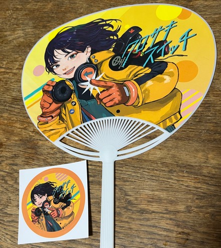 クロサキスイッチのロゴがデザインされたカラフルなうちわとステッカーが並べられている写真。うちわにはイベント名とイラストが描かれ、ステッカーは丸型や四角型など複数の形があり、来場者が記念に持ち帰る姿も見られる。黒崎の街のにぎわいを象徴するアイテムとして、テーブルの上に明るく映えている。